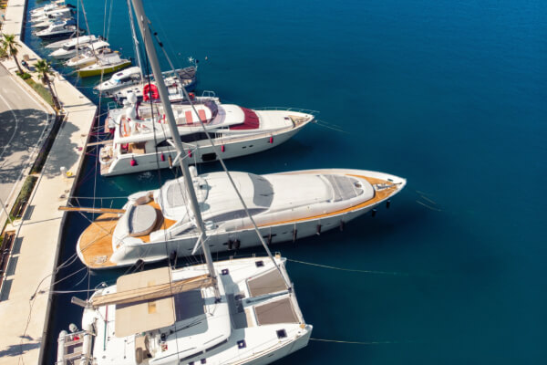 An aerial view of a clean marina with a concrete pier where multiple luxury boats, including white yachts and catamarans, are docked on a sunny day with clear water.