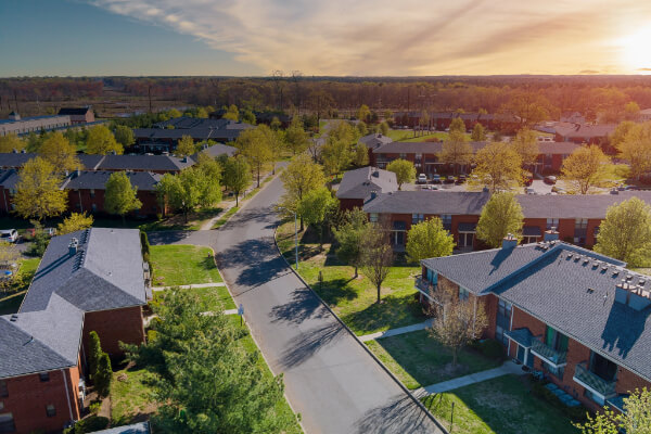 An aerial view of a peaceful residential suburban neighborhood with rows of houses and lush green lawns. The winding road is surrounded by trees, with a forest in the background under a sunset sky.