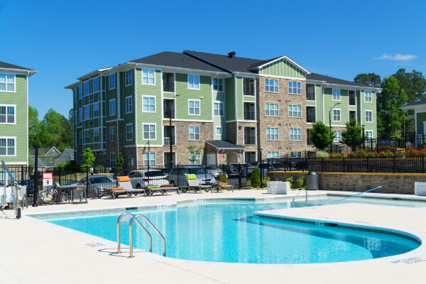 An outdoor swimming pool with a shallow end, built for an apartment complex with green-sided residential buildings in the background. The area includes lounge chairs and a security fence.