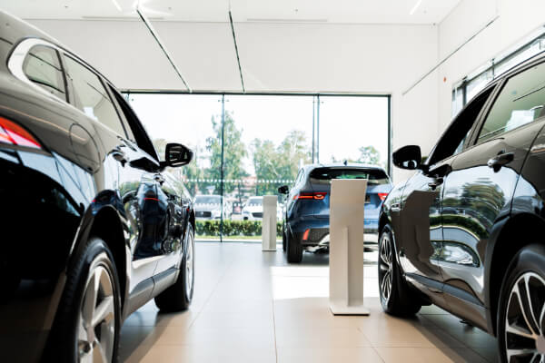 A wide-angle, hyper-realistic, and brightly lit view from inside a modern automotive group showroom. Two new black SUVs are visible in the foreground with another dark-blue car in the background, all on display for sale, showcasing the clean, minimalist interior with large windows.