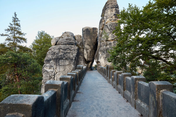 A wide-angle, low-angle shot of the Bastei Bridge, a famous stone bridge in Saxon Switzerland National Park, Germany. The bridge is surrounded by tall, natural rock formations, and the walkway is lined with a low stone fence.