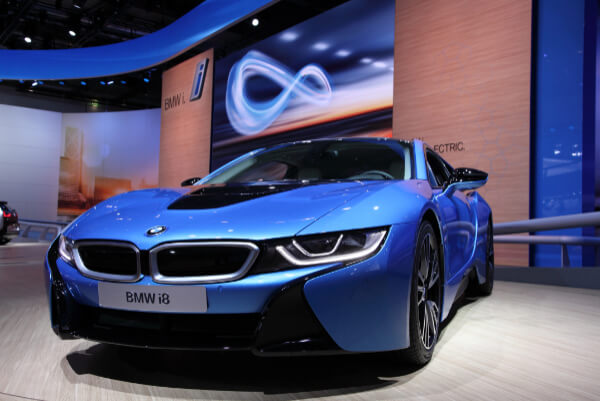 A front-angle, low-angle shot of a blue BMW i8 sports car with black accents on a white platform inside a brightly lit dealership or auto show. The vehicle has a prominent front grille and a large screen with a logo in the background.