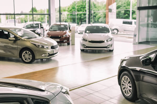 A wide-angle, low-angle, and slightly blurry shot of a car dealership showroom floor, with multiple new and used vehicles on display. The clean, modern interior has a polished floor and a large glass window showing a street in the background.