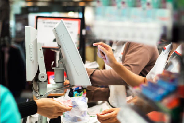 A close-up shot of a cashier at a brick-and-mortar retail store checkout counter, with their hand reaching toward a customer's hand, who is holding a stack of money. The scene is slightly blurry, with a POS system and other staff in the background.