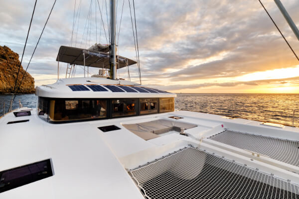 A view from the deck of a luxury catamaran with solar panels on the roof and trampolines on the bow. The image is taken at sunset, with a golden sky and calm ocean in the background. The mast and rigging are visible against the sky.