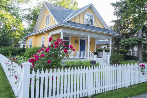 A charming, yellow, two-story custom-built house with a dark blue front door and a covered porch. A well-maintained garden with vibrant pink roses and green bushes is in the foreground, enclosed by a classic white picket fence.