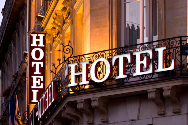 A low-angle shot of a classic European hotel facade at dusk, featuring two prominent, illuminated "HOTEL" signs with bold, white letters and a red background, set against an ornate iron balcony railing.