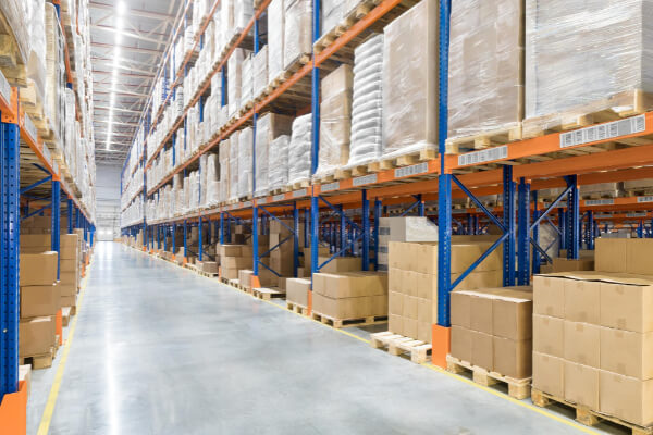 A wide-angle view of a clean, well-lit modern warehouse showing an aisle between tall metal racks filled with goods, including cardboard boxes and shrink-wrapped pallets.