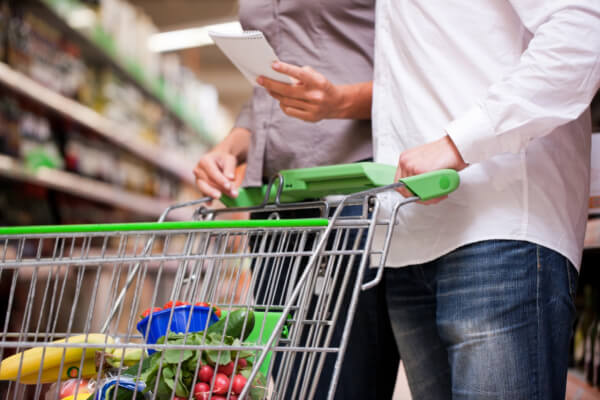 A couple is grocery shopping in a brick-and-mortar retail store. The woman holds a shopping list, and the man pushes a shopping cart filled with fresh produce, with aisles and shelves of products in the blurry background.