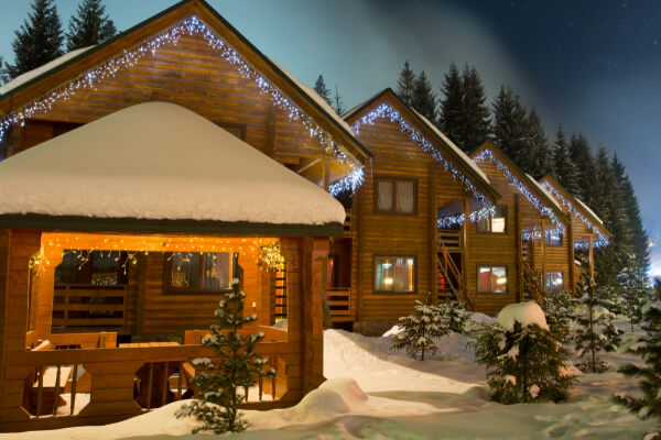 A row of cozy wooden chalets covered in snow and decorated with festive string lights at night. The properties are surrounded by snow-covered pine trees under a starry winter sky, with a lit-up gazebo in the foreground.