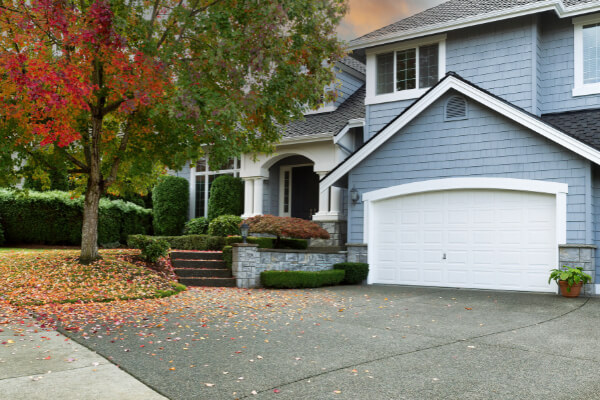 A large two-story custom-built house with gray siding, a prominent white garage door, and a covered front porch with white pillars. The front yard is covered with colorful fallen autumn leaves, and the landscaping includes a stone wall, concrete steps, and a decorative tree with red and green foliage.