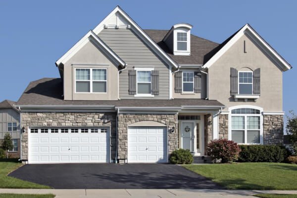 A large, custom-built, two-story house featuring a mix of gray siding, light-colored stucco, and a prominent stone facade on the lower level. The home has a gabled roof, a two-car garage with white doors, and a well-kept front yard.