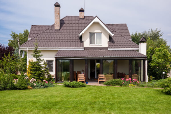 A two-story custom-built house with a gabled brown shingle roof, beige siding, and a large covered porch area with wooden pillars. The front yard has a well-maintained green lawn, mature trees, and flowering bushes, with a paved path leading to the entrance.