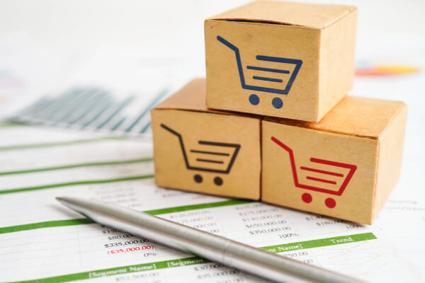 A close-up, high-angle shot of three miniature cardboard boxes with shopping cart logos stacked on top of each other. The boxes are placed on a financial spreadsheet with a silver pen, representing a business concept for e-commerce.