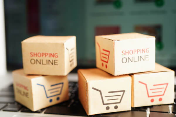 A close-up, eye-level shot of several small cardboard boxes with the words "Shopping Online" and a shopping cart logo printed on them. The boxes are stacked on a laptop keyboard, with a website visible on the screen in the background.