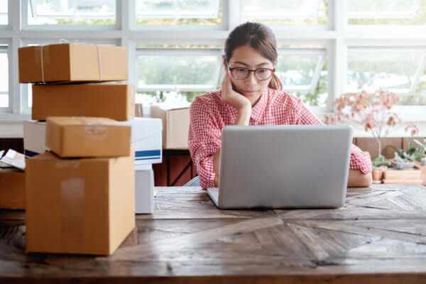 A young female entrepreneur wearing glasses and a plaid shirt, working on a laptop at a wooden table. She is surrounded by a stack of cardboard boxes with various shipping labels, suggesting she is managing an e-commerce business or fulfilling online orders.