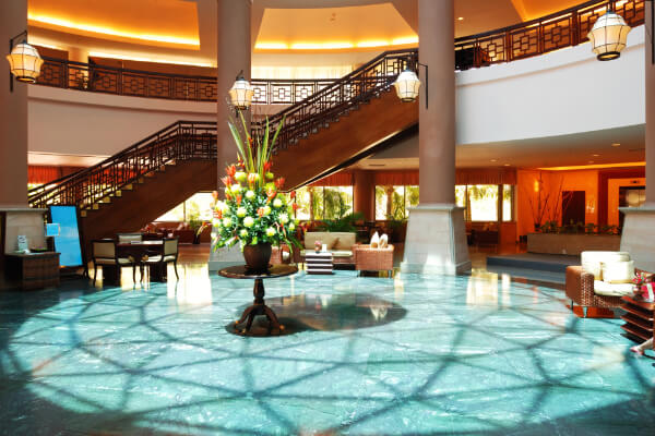 A grand, upscale hotel lobby with a large floral arrangement on a round table in the center, a sweeping staircase leading to a second floor, and various seating areas.