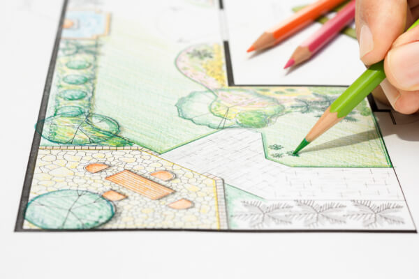 A close-up of a hand using a green colored pencil to sketch and color a detailed landscape design plan. The blueprint includes a stone patio with outdoor furniture, various plants, and a lawn area.