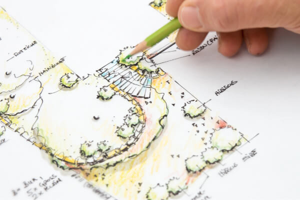 A detailed, hand-drawn landscape design plan for a residential garden, featuring a patio, plants like hibiscus and roses, and a lawn area. A hand is visible holding a green pencil, actively coloring the blueprint.