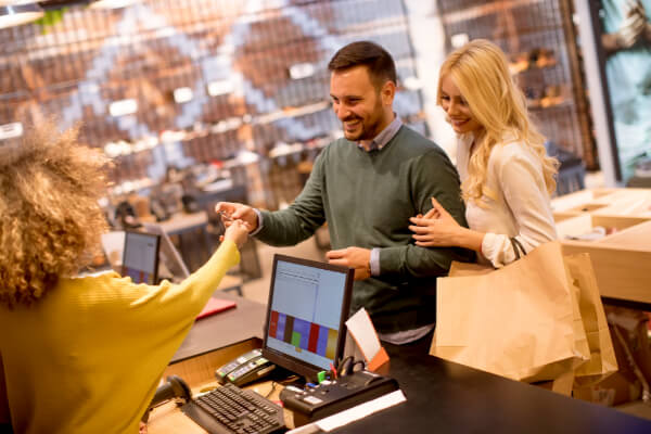 A wide-angle shot of a happy young couple in a brick-and-mortar retail store, making a payment at the checkout counter. The man is handing a credit card to the cashier, while the woman holds a shopping bag and smiles. The background shows retail shelves and clothing displays.