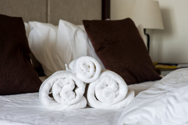 Close-up of three freshly laundered, fluffy white towels neatly rolled and stacked on top of a soft hotel bed with crisp white linens and brown pillows.