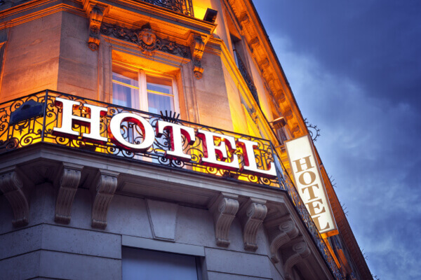 A close-up, low-angle shot of a classic stone hotel building at dusk with a large, illuminated "HOTEL" sign in glowing white and red letters, mounted on an ornate balcony railing.