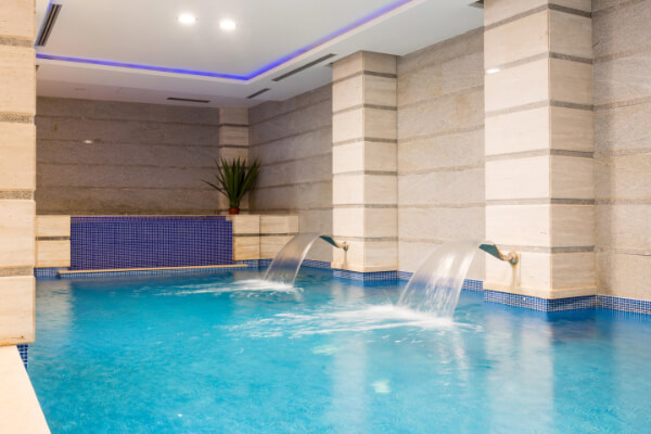 A view of a modern indoor hydrotherapy pool with blue mosaic tiles, two wall-mounted waterfall jets, and textured stone walls under soft ambient lighting, creating a relaxing wellness atmosphere.