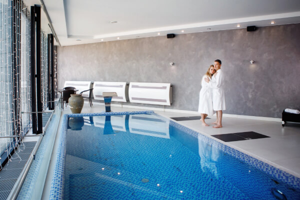 A couple in white bathrobes stands next to a small indoor swimming pool with blue tiles. The pool is in a modern, well-lit room with floor-to-ceiling windows and a gray textured wall.