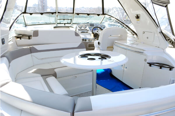 A wide-angle view of the pristine interior cockpit of a luxury powerboat, showing white cushioned seating with gray trim, a small round table, and a steering wheel at the helm. The boat is on the water on a sunny day.
