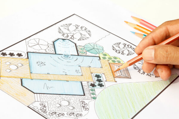 A detailed, hand-drawn landscape architecture blueprint and design plan of a residential backyard with a large swimming pool, a deck, a stone patio, and various trees. A hand is visible holding an orange colored pencil, actively coloring the plan.