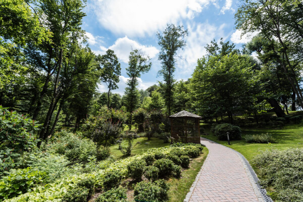 A wide-angle, hyper-realistic view of a beautifully designed park or residential landscape with a winding red brick path leading toward a small wooden gazebo. The area is surrounded by lush green trees, shrubs, and a clear blue sky with white clouds in the background.