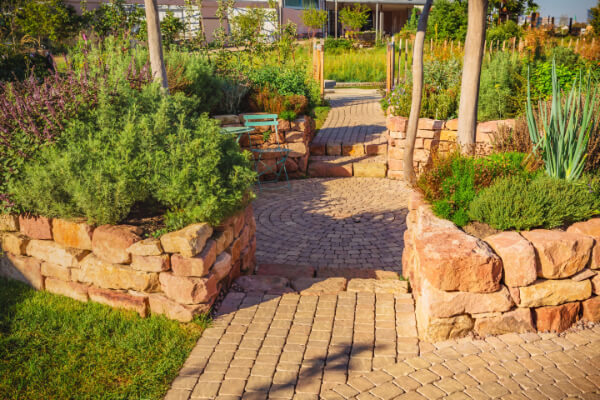 A vibrant, sunny image of a lush backyard garden with a variety of green plants and a winding, light-brown cobblestone path. The path is bordered by low walls made of natural red stones, with a small seating area and a circular stone pattern in the center.