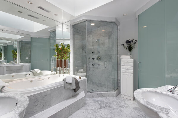 A luxurious, spacious bathroom with white and gray marble tiles on the floor and bathtub surround. The room features a large whirlpool tub, a glass-enclosed shower with multiple jets, and a large mirror reflecting the space.