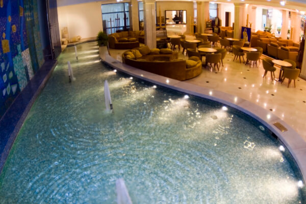 A wide-angle view of a luxurious hotel lobby featuring a large, curved indoor fountain with multiple water jets and a seating area with gold velvet couches and round tables.