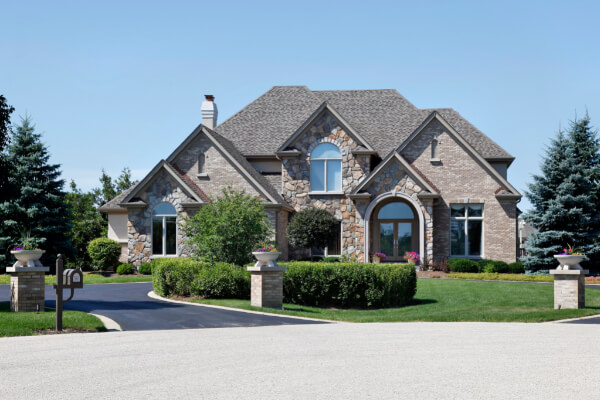 A large, luxurious two-story residential house featuring a facade of natural stone and brick with a gabled shingle roof. A well-maintained green lawn, manicured hedges, and a paved driveway lead to the house, flanked by tall pine trees and decorative mailboxes.