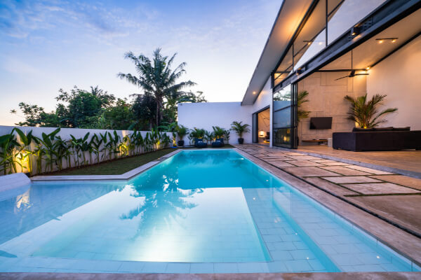 A serene backyard view of a luxury modern villa with a clear blue swimming pool at dusk. The house features floor-to-ceiling glass doors that open to a patio with stone pavers, and the pool area is surrounded by lush green plants and palm trees.