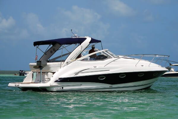 A wide-angle, side-view shot of a white and navy blue luxury motor yacht on light blue water with two people in the cockpit. The boat has a blue canvas bimini top, a swim platform, and a sunbathing area in the front, with a clear sky in the background.