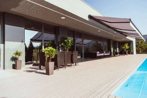 A view of the spacious wooden deck and outdoor seating area of a luxury residential villa. The modern house has large floor-to-ceiling glass doors, a large awning for shade, and a clean, light-blue swimming pool next to the deck.