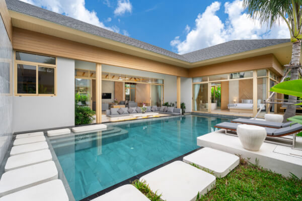 A view of a luxurious residential villa with an indoor-outdoor swimming pool and a light gray marble interior. The house has floor-to-ceiling glass doors, a patio with lounge chairs, and a lush lawn surrounded by a white wall.