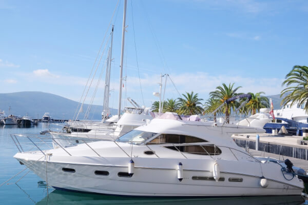 A wide-angle, sun-drenched shot of a luxurious white motor yacht docked in a tropical marina. Other boats and sailboats are docked nearby, with palm trees and a mountain in the background against a clear blue sky.