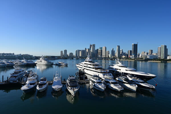 An aerial wide-angle shot of a sunny day at a Miami marina, with many luxury yachts and boats of various sizes docked in the calm blue water. In the background, the city's skyline with tall skyscrapers is visible against a clear blue sky.