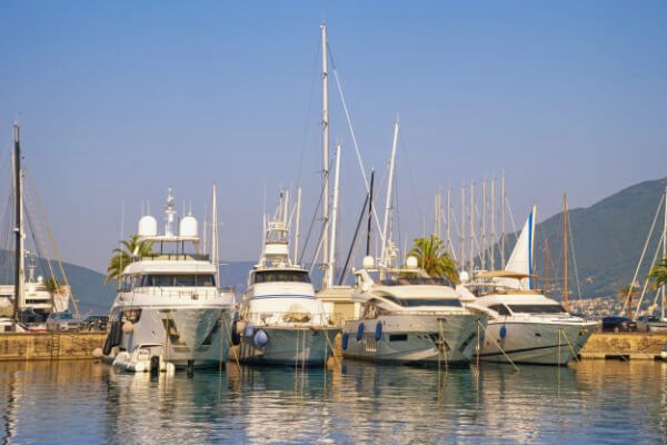 A wide-angle, sun-drenched shot of several luxury yachts and large motorboats of various sizes docked in a marina on a calm bay. The water reflects the boats, and the background shows a clear blue sky, green hills, and a port with other boats and waterfront buildings.
