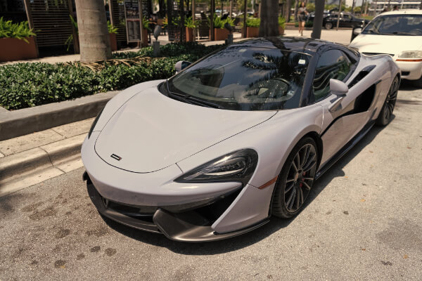 A front-angle, close-up view of a parked McLaren 570S luxury sports car with a sleek, light gray finish and a black carbon fiber roof. The vehicle is on a paved street next to a sidewalk with lush green plants and palm trees.