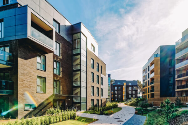 A wide-angle, sun-drenched view of a modern apartment or condominium complex with several multi-story buildings. The residential properties have a mix of brick and dark siding with large windows and glass balconies. The grounds are well-landscaped with green lawns and paved walkways.