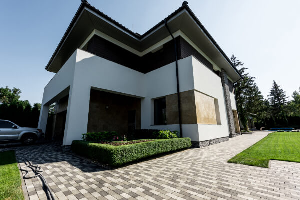 A modern custom-built, two-story house with a flat roof, white and brown siding, and stone accents. The front yard features a well-manicured hedge in a raised garden bed, a block-paved driveway, and a small patch of green lawn. A silver SUV is visible in the garage.