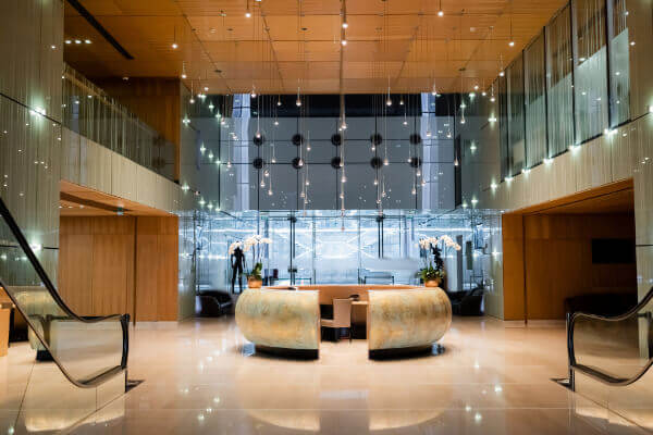 A view of a sophisticated and modern hotel lobby featuring a unique, segmented reception desk with a textured, metallic finish, surrounded by an open atrium with a glass wall and suspended light fixtures.