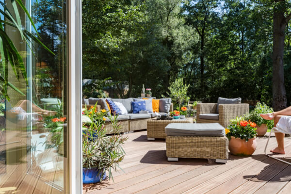 A wide-angle, hyper-realistic view of a spacious wooden garden deck with a complete set of modern, woven rattan outdoor furniture. The deck is surrounded by lush greenery, potted plants, and trees, with a large sliding glass door reflecting the interior of the house.