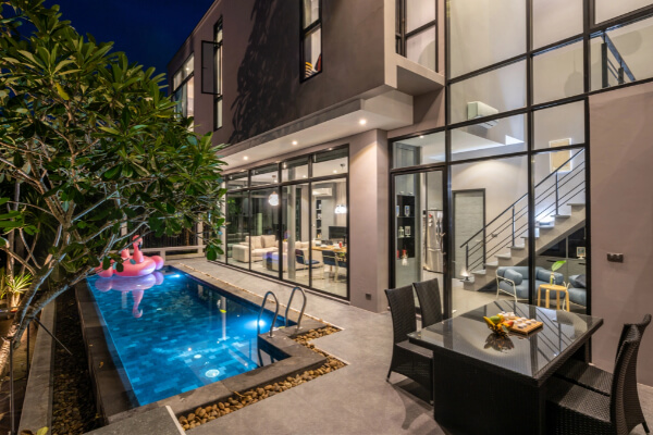 A modern residential house at night with an in-ground, rectangle swimming pool and a pink inflatable flamingo floating inside. The pool area includes an outdoor dining table with chairs and is surrounded by lush tropical plants. The house has large floor-to-ceiling windows showing a bright interior.