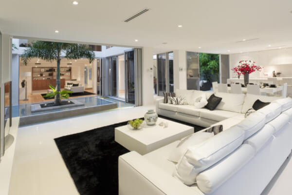 A wide-angle, hyper-realistic, and brightly lit image of a modern residential home interior with an open-concept living room. The space has white tile floors, a large white sectional sofa, a black rug, and a small, square, indoor courtyard with a palm tree in the center.