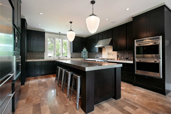 A wide-angle view of a modern kitchen with dark black wooden cabinets, a large island with a stainless steel countertop and four industrial-style stools. The kitchen features stainless steel appliances, a unique tile backsplash, and a light-brown marble tile floor.